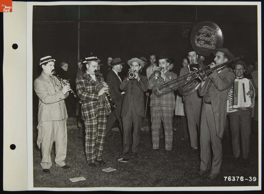 Group of seven men in suits play instruments on a field with people in the background