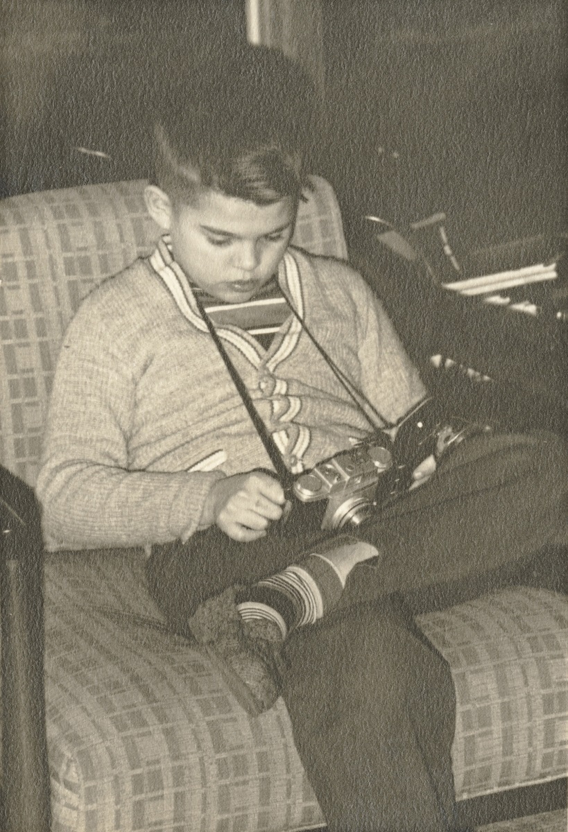 Black and white image of young boy sitting on chair looking at camera on a strap around his neck