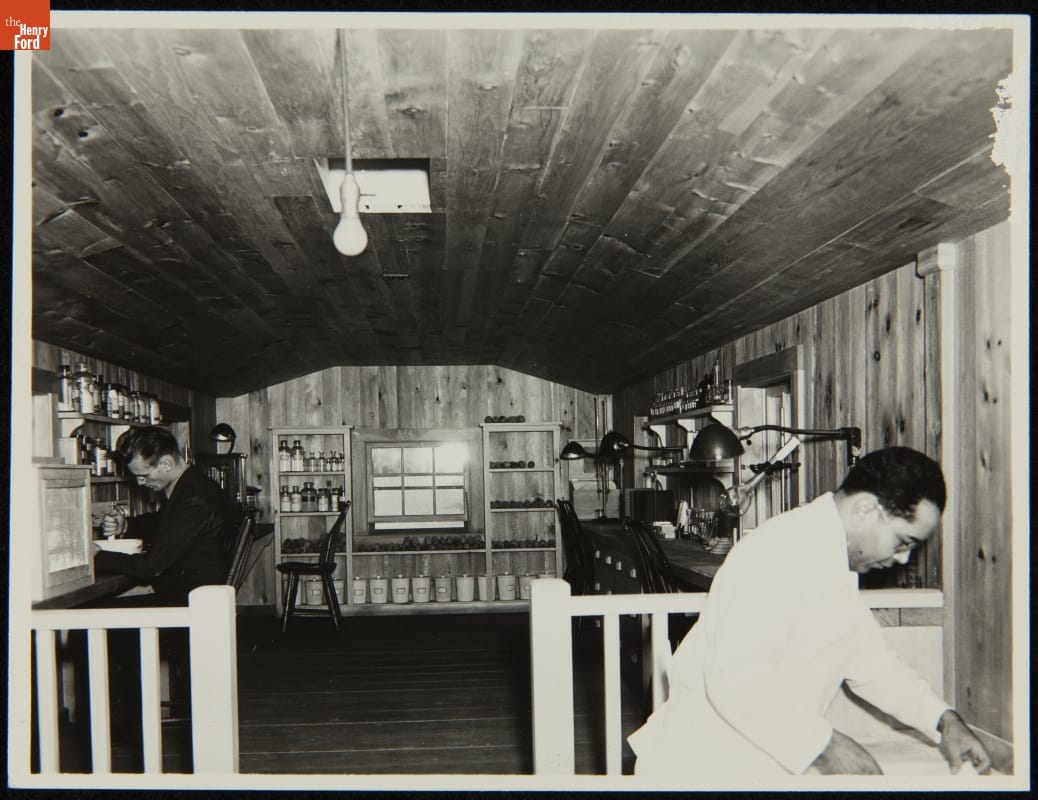 Room with work surfaces along walls filled with equipment; two men work in the space