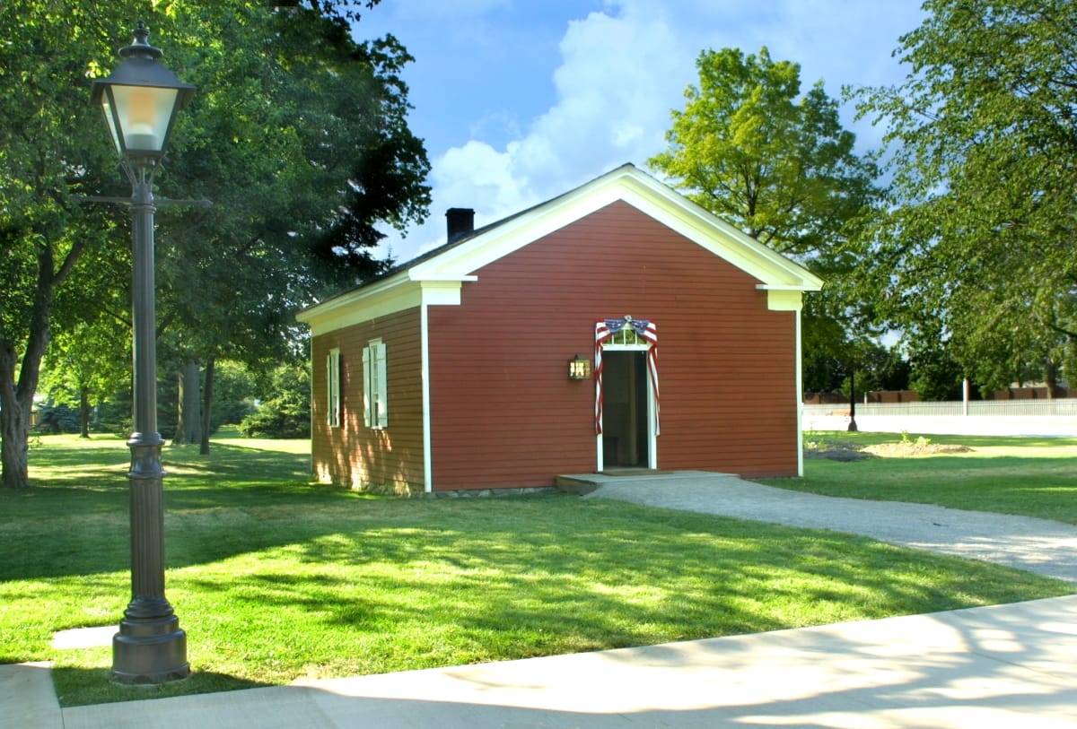 Small red wooden building among green lawns, trees, walkways, and streetlamps