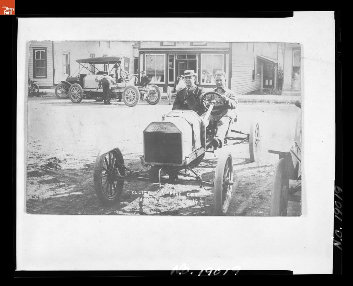 Black-and-white photo of two men sitting in an early open car on a street with other cars, people, and buildings in the background