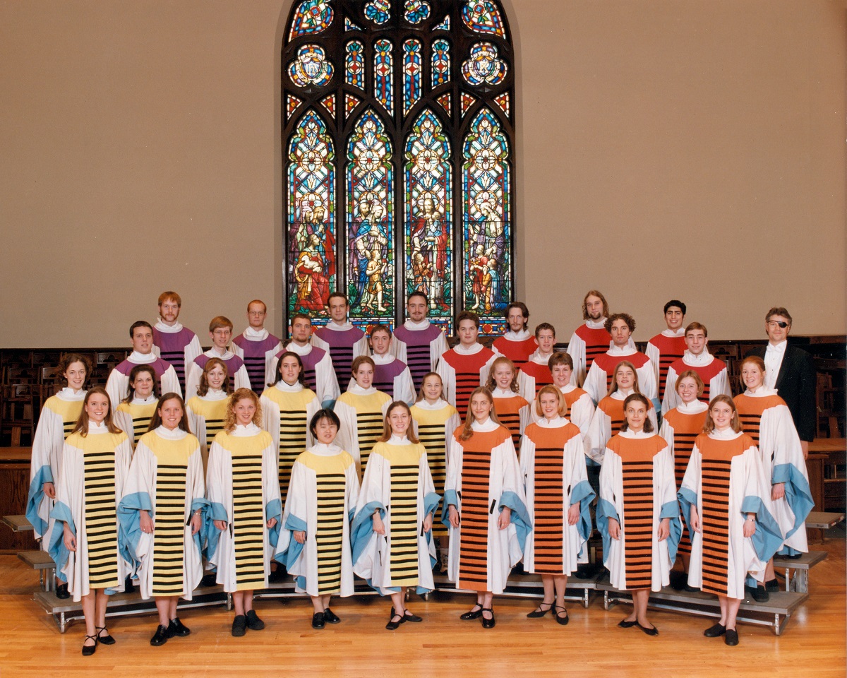 Group of people in colorful robes with black stripes, standing on bleachers in front of a stained glass window 