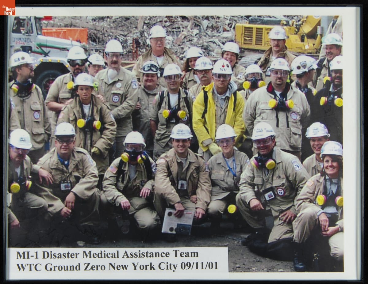 Group of people, mostly in hard hats and khaki clothing, many with respirators, pose in front of a pile of rubble and construction equipment