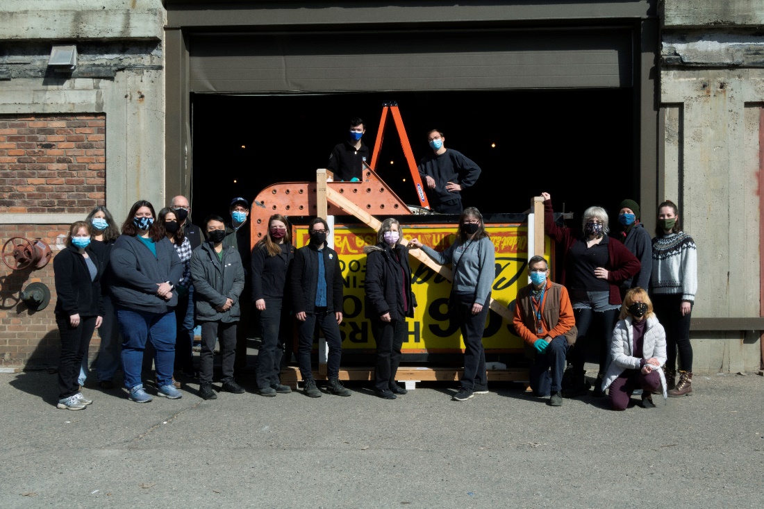 A group of people wearing masks pose for a photograph in front of, and behind, on a ladder, a large sign