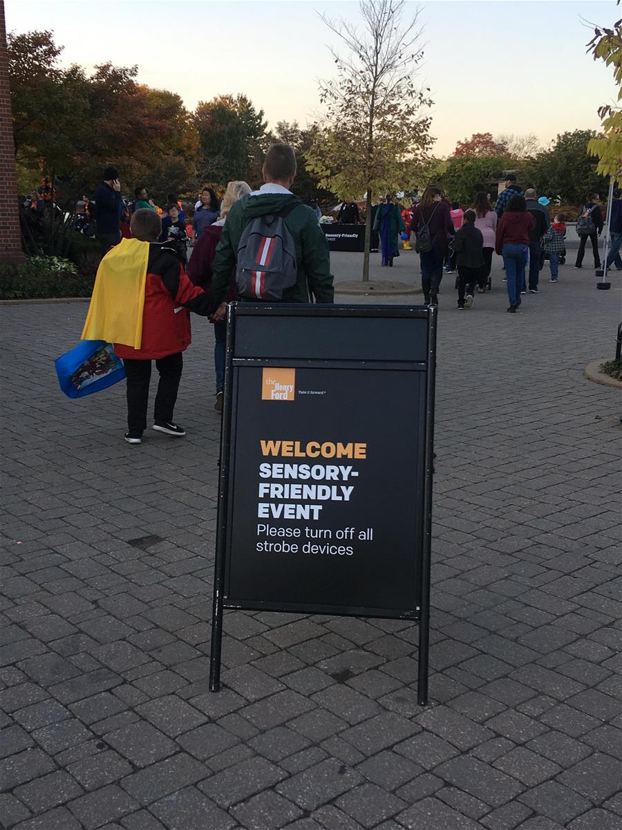 Sign with text in foreground; plaza with many people and trees behind