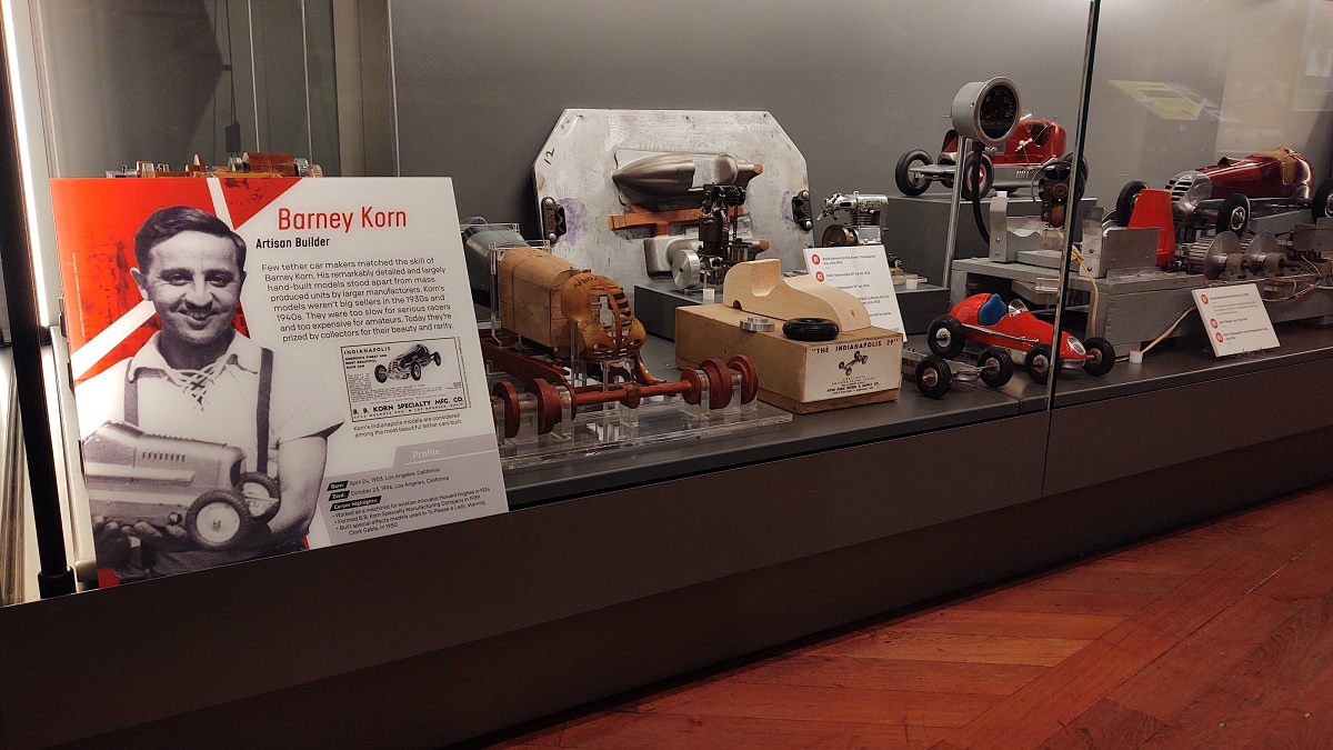 Glass display case containing a sign with text and images and model cars, both whole and in parts and pieces