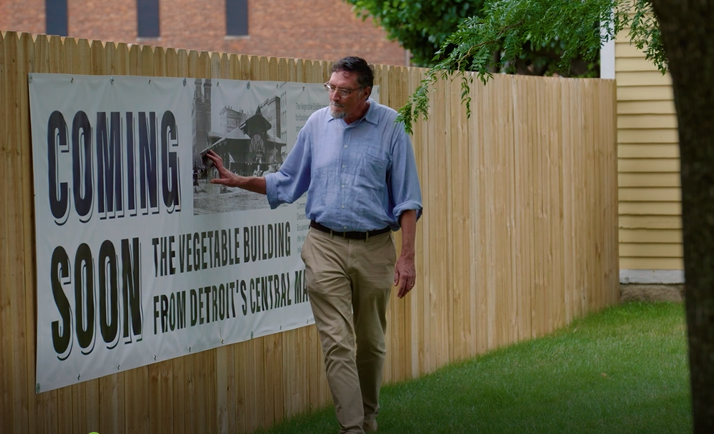 Greenfield Village Director Jim Johnson walks along the fence around the Detroit Central Farmers Market construction site in Greenfield Village Man in blue button-down shirt and khaki pants walks along wooden fence with large white banner reading
