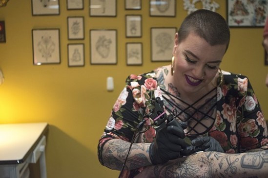 Carrie Metz-Caporusso Smiling person in black gloves tattoos another person's arm; framed art hangs on a wall behind them
