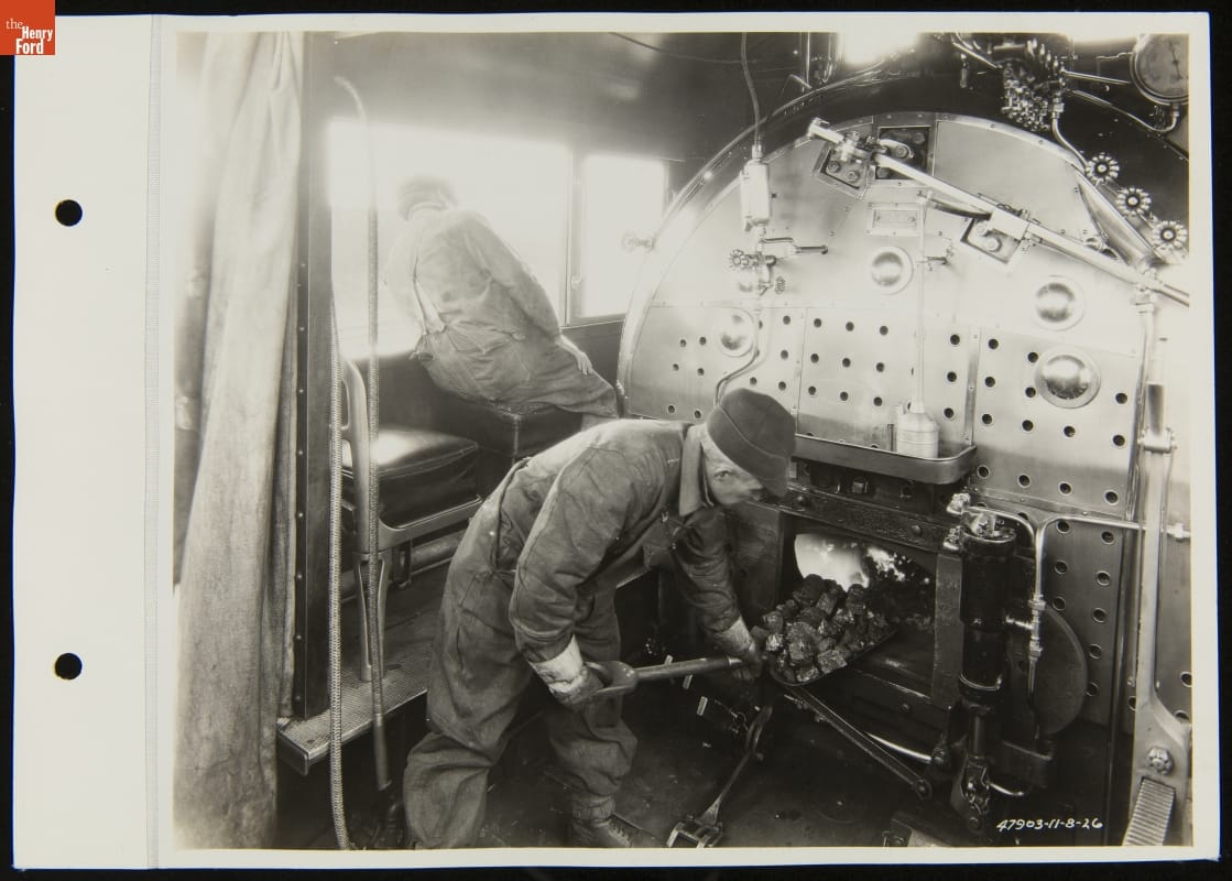 Black-and-white image of two men, one shoveling coal into a large metal furnace and the other leaning out a window