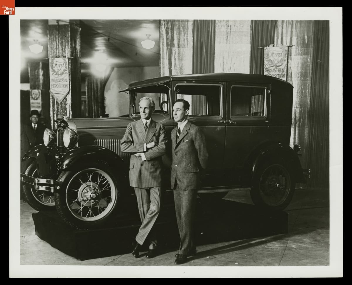 Two men wearing suits stand by car in indoor space with lights and banners